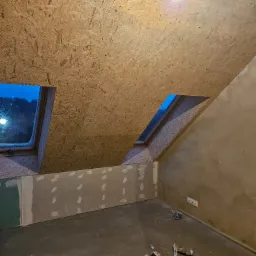 Poddasze w trakcie wykańczania z oknami dachowymi i płytami OSB na suficie, widoczne niedoskonałości na ścianach i narzędzia na podłodze.