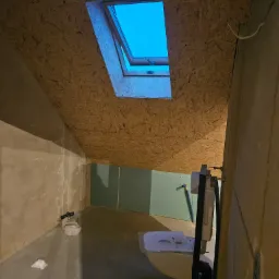 Surowe wnętrze poddasza z oknem dachowym i częściowo zamontowaną ścianką działową z zielonych płyt gipsowo-kartonowych, widoczna instalacja sanitarna i fragment nieotynkowanej ściany.