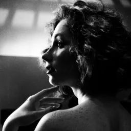 Czarno-biały portret studyjny kobiety z kręconymi włosami, profil, z ręką pod brodą, widoczne piegi na ramionach, oświetlenie padające z góry tworzące geometryczne cienie na ścianie w tle.