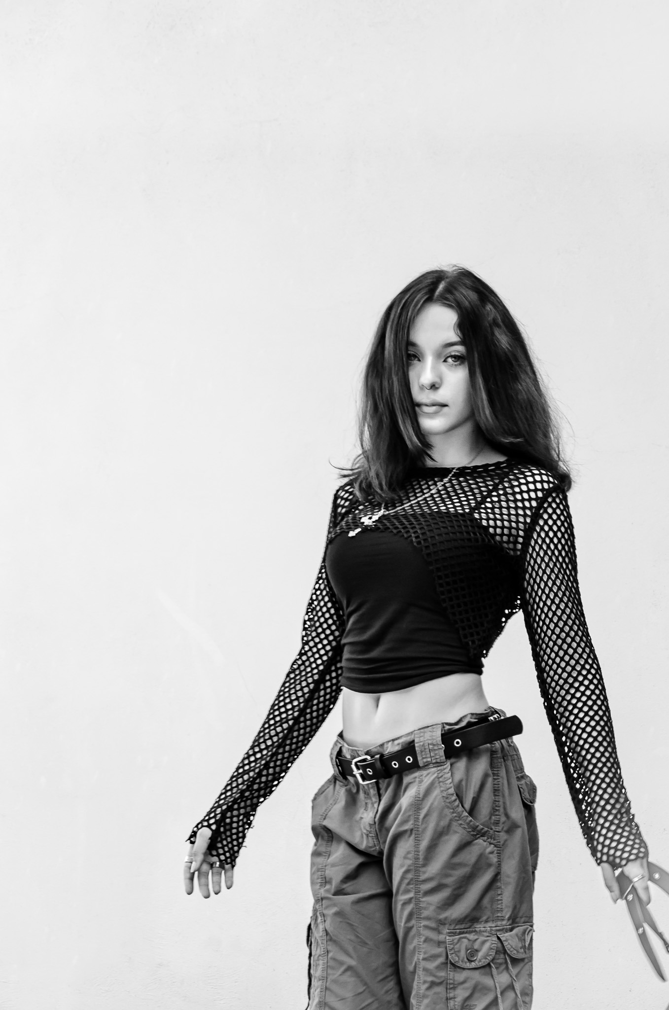 Czarno-białe zdjęcie młodej kobiety w siateczkowej bluzce, topie i spodniach cargo, pozującej na białym tle. Widoczny piercing w nosie i srebrny naszyjnik.