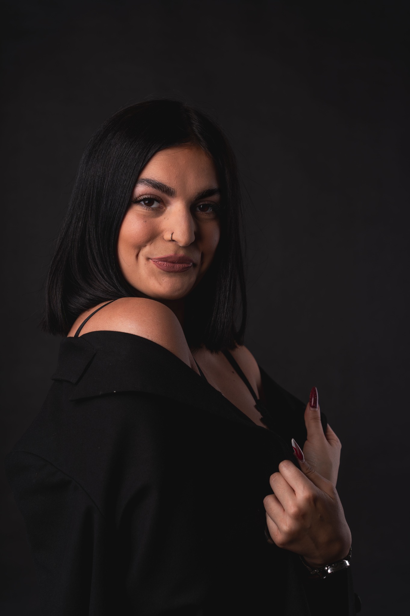 Portret uśmiechniętej kobiety z kolczykiem w nosie, ubrana w czarny żakiet z odsłoniętym ramieniem, na ciemnym tle.