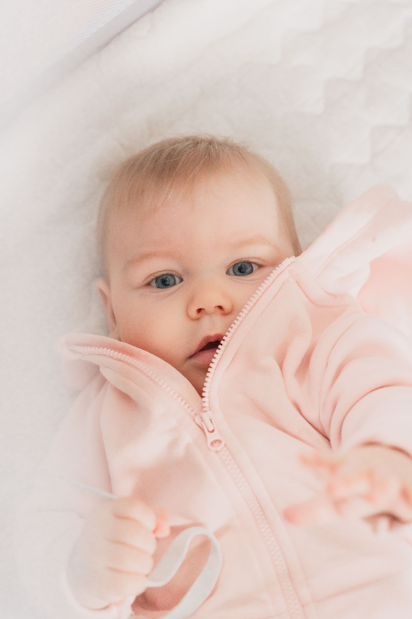 Niemowlę z blond włosami i niebieskimi oczami leży na białym kocyku, ubrane w różowy kombinezon z rozpiętym zamkiem błyskawicznym.
