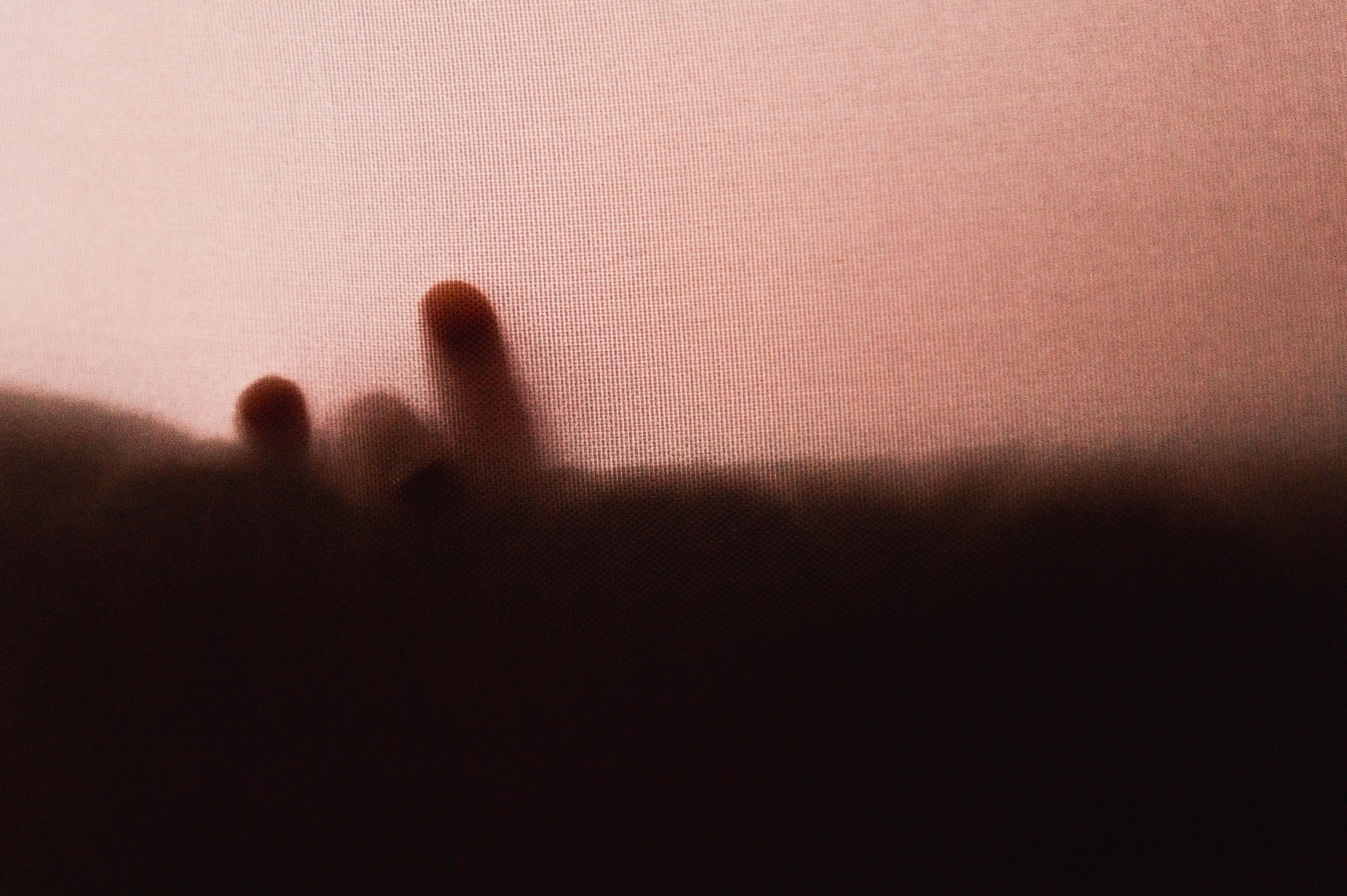 Delikatne zarysy dłoni noworodka, widoczne przez cienką, różową tkaninę o wyraźnej fakturze, tworzące subtelny, rozmyty obraz.