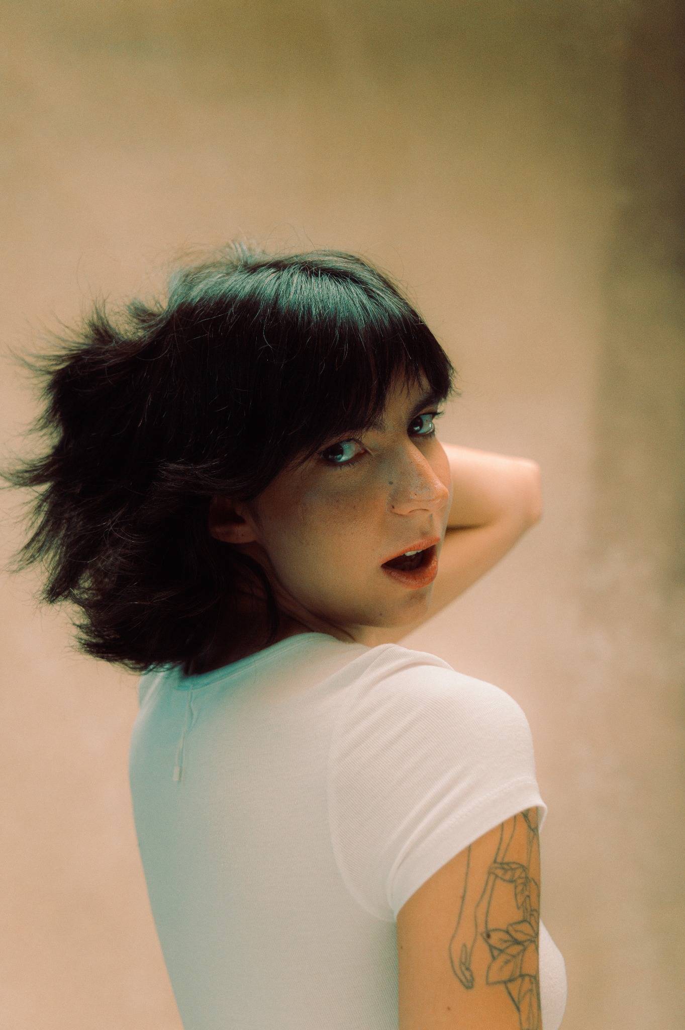 Portret kobiety z krótkimi, ciemnymi włosami, odwracającej się przez ramię, w białej koszulce z tatuażem na ramieniu, na rozmytym beżowym tle.