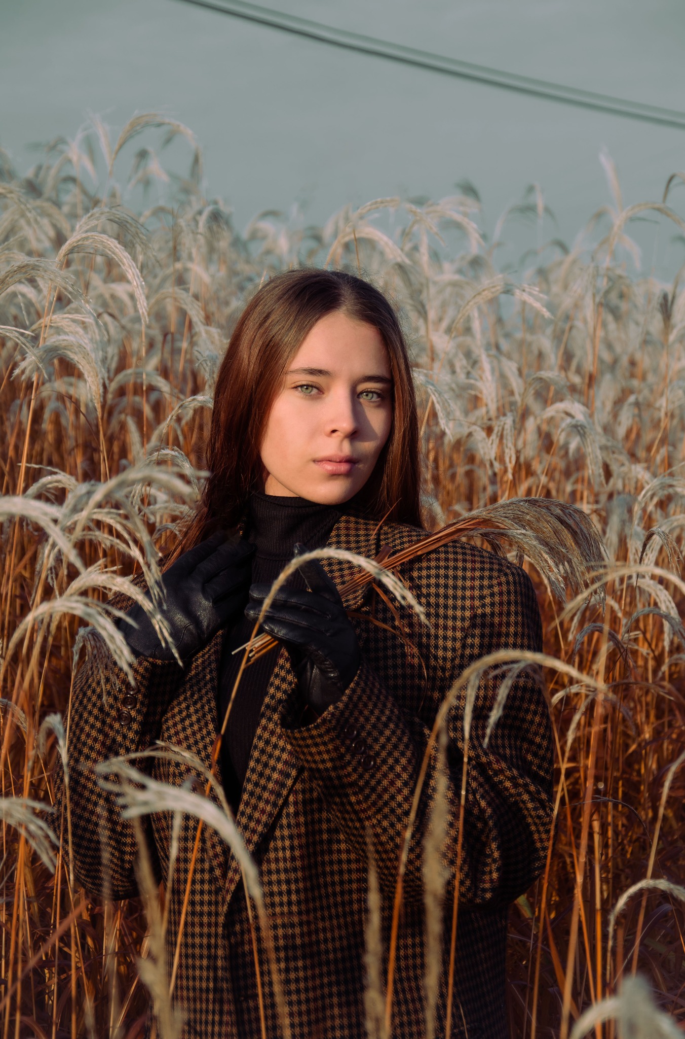 Portret kobiety w brązowo-czarnej kraciastej marynarce i czarnych skórzanych rękawicach, stojącej w plenerze pośród wysokich traw z pióropuszami, trzymającej w dłoniach garść tych traw.