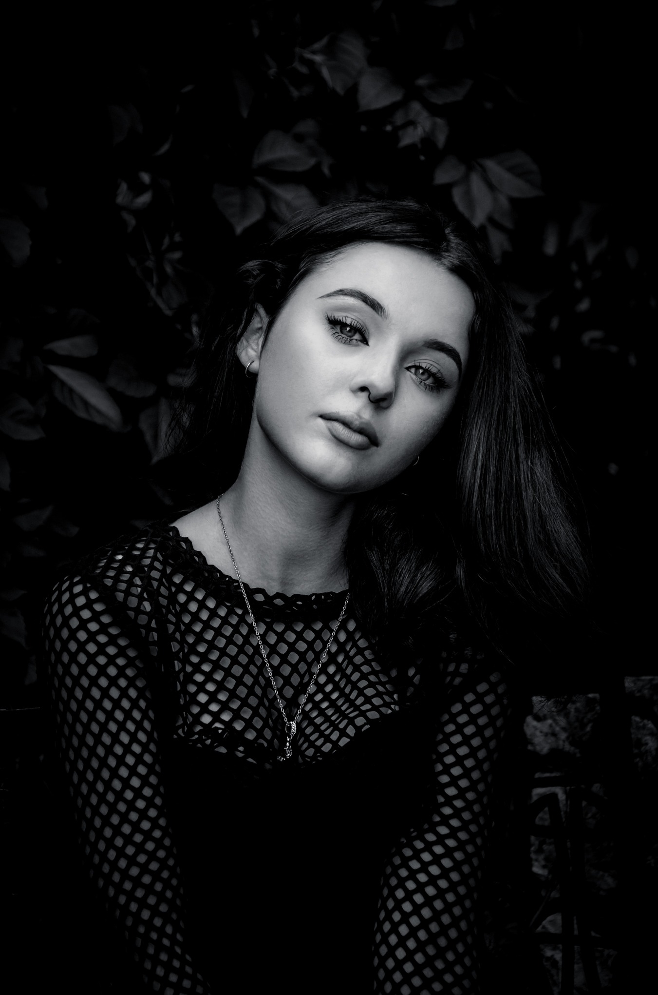 Portret młodej kobiety w czarno-białej tonacji, z delikatnym makijażem i srebrnym naszyjnikiem na tle ciemnych liści, ubrana w siatkową bluzkę.