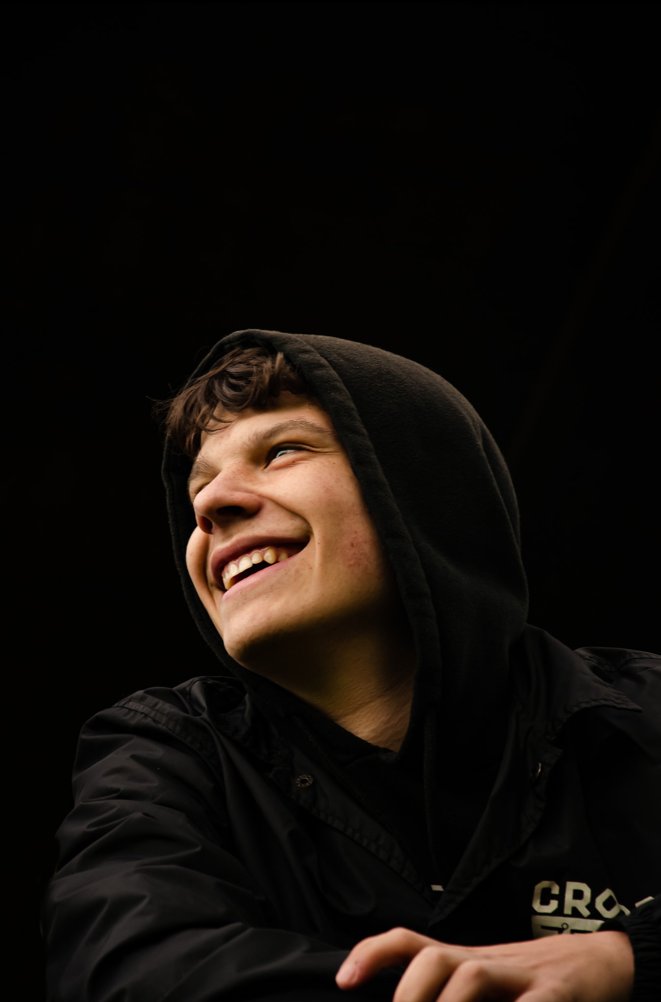 Młody mężczyzna w czarnej bluzie z kapturem, uśmiechający się i patrzący w górę, sfotografowany w Tarnowie na ciemnym tle.