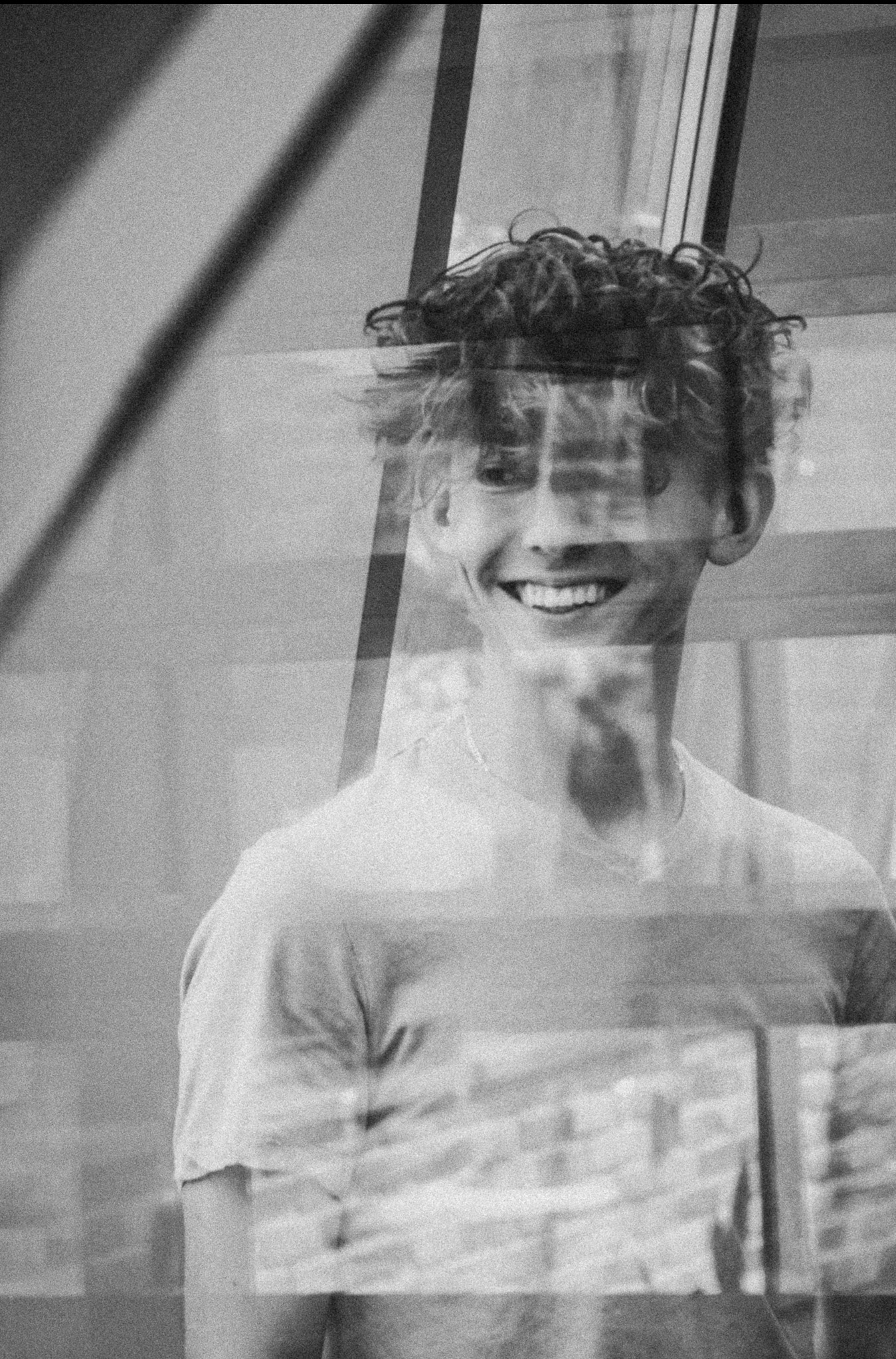 Portret młodego mężczyzny z kręconymi włosami, uśmiechającego się i patrzącego w bok, z efektem nałożonych warstw i odbić, w czerni i bieli.