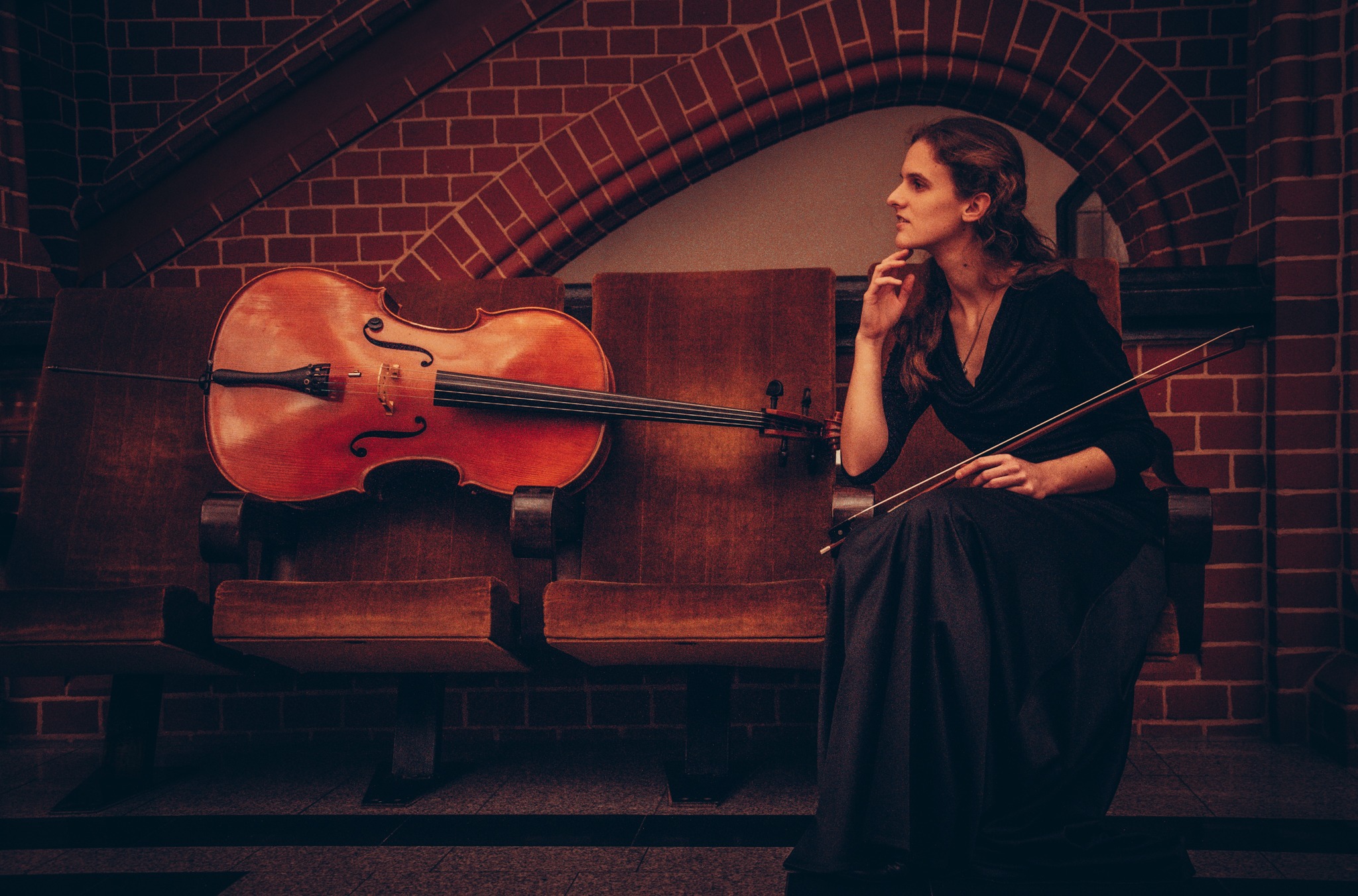 Kobieta w czarnej sukni siedzi na drewnianym krześle, trzyma smyczek; obok wiolonczela oparta o krzesło, w tle ceglany łuk.