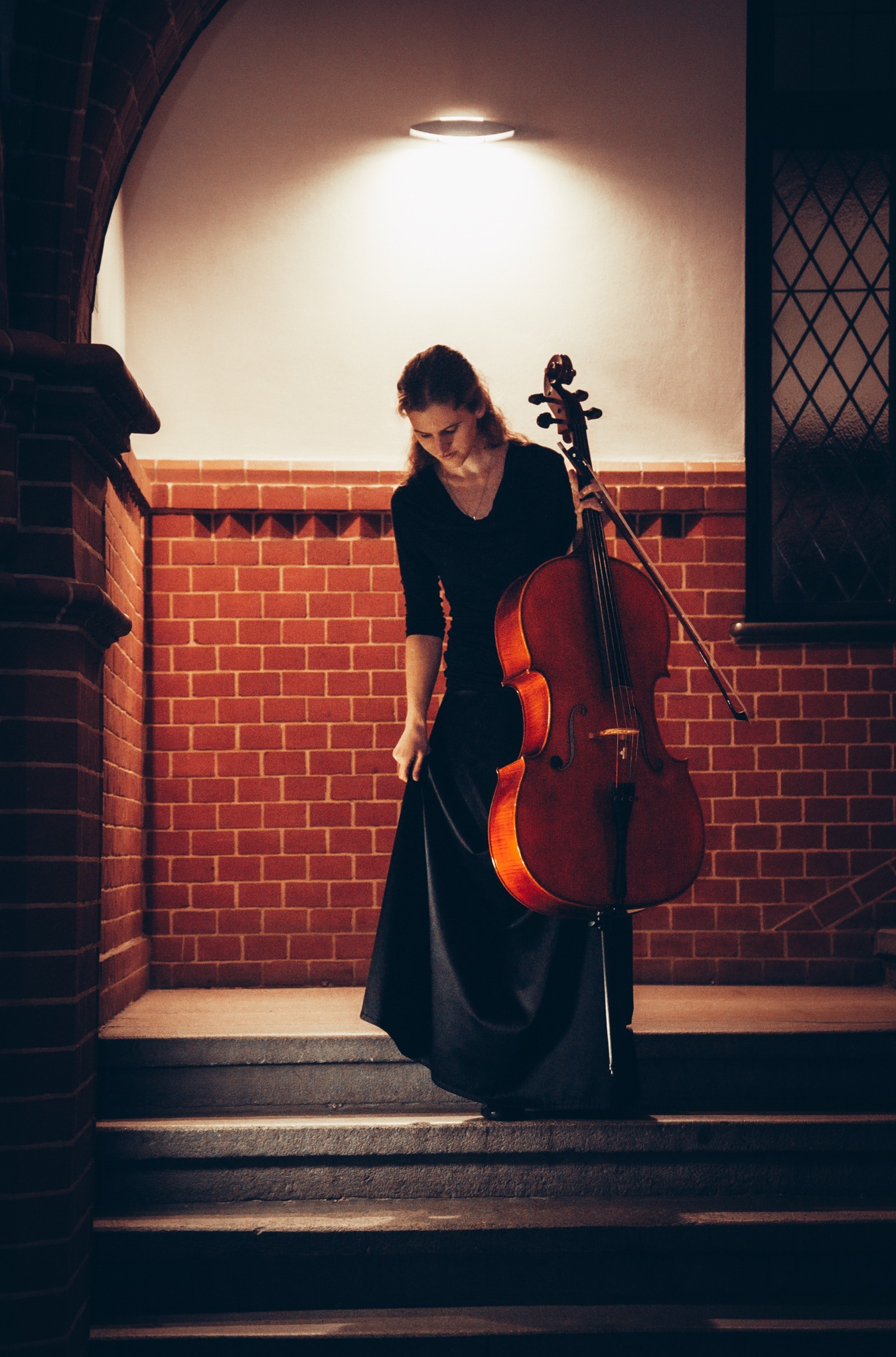 Kobieta w długiej czarnej sukni trzyma wiolonczelę na tle ceglanej ściany i okna z kratami, stojąc na schodach.