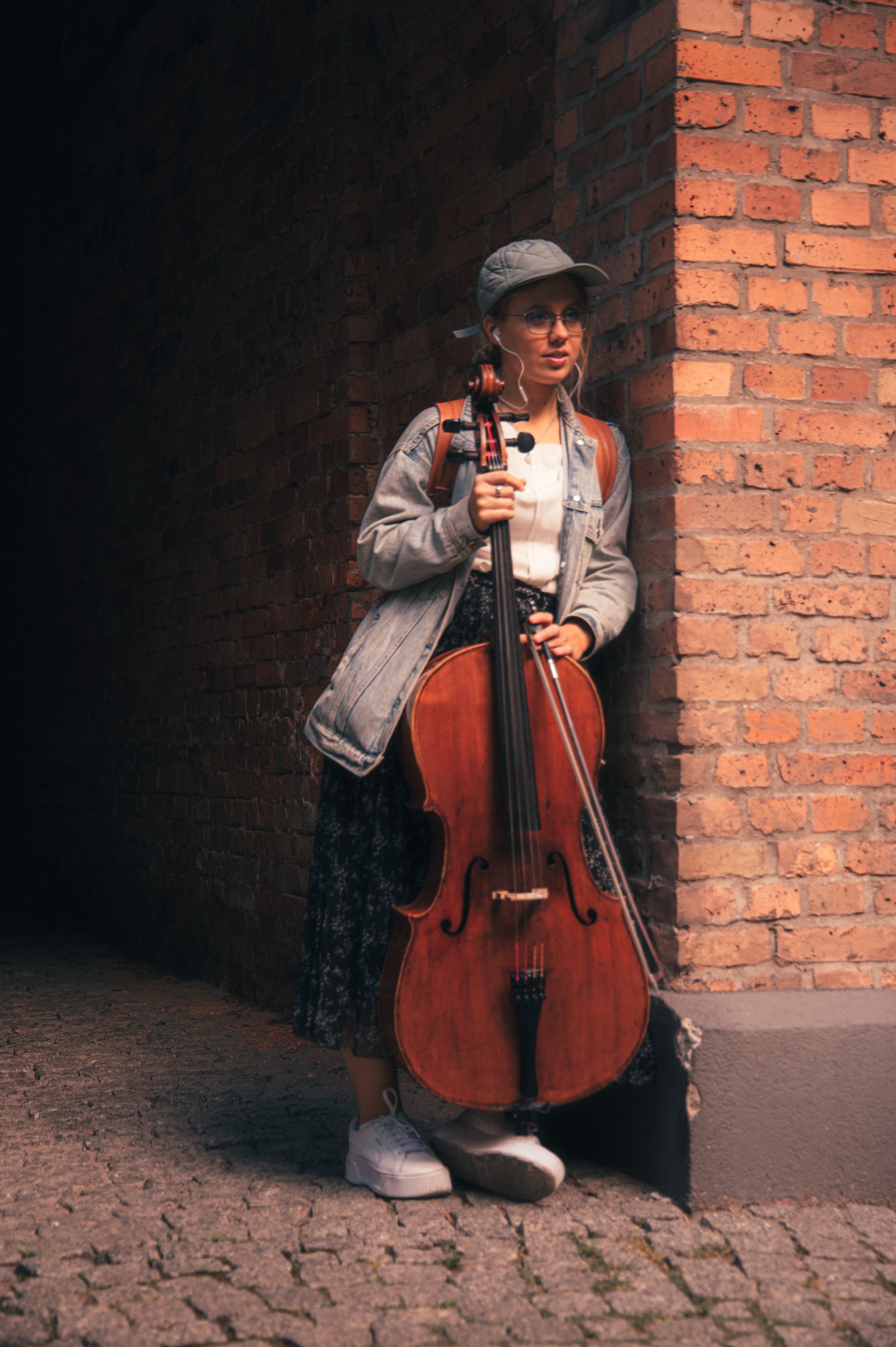 Kobieta w czapce i okularach opiera się o ceglany mur, trzymając wiolonczelę w wąskim przejściu, oświetlonym ciepłym światłem.
