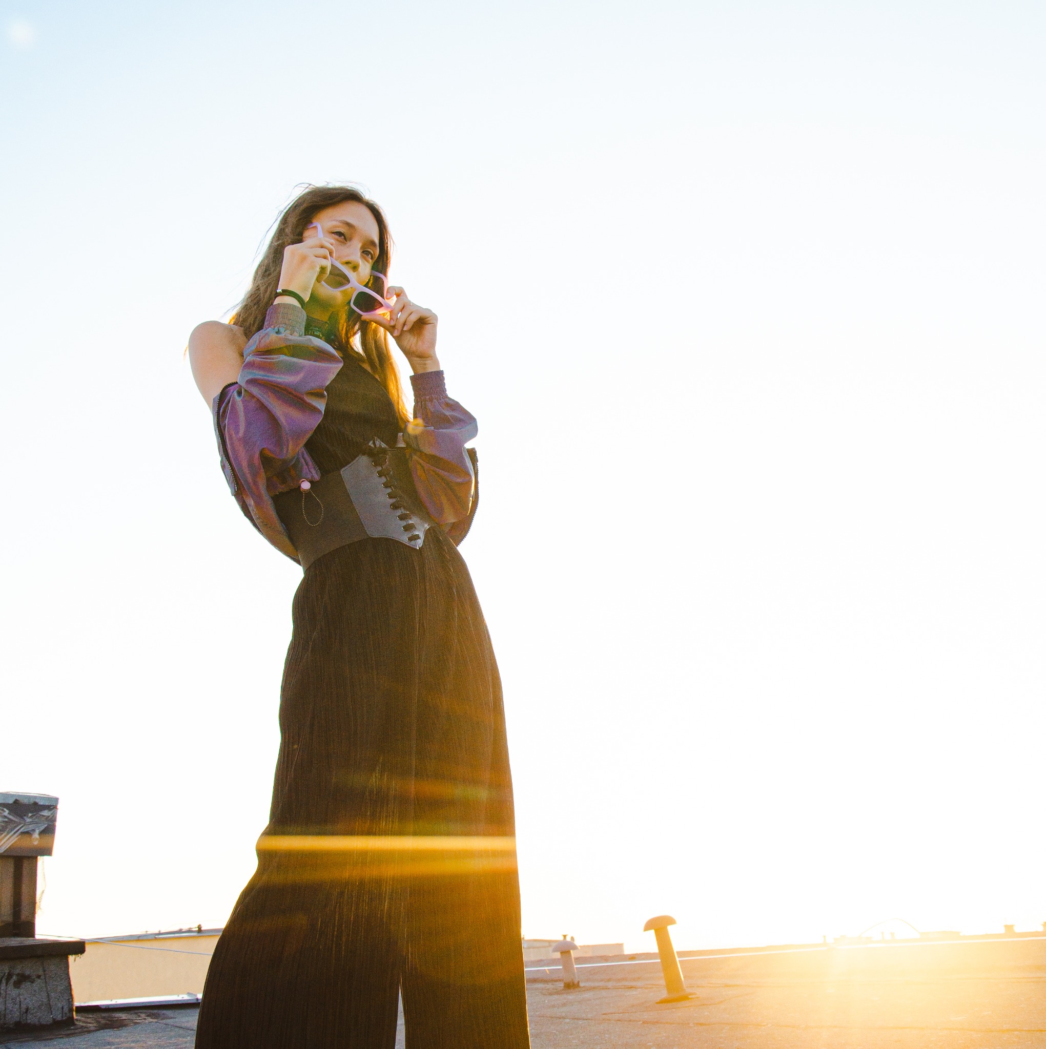 Kobieta w szerokich spodniach i bluzce z bufiastymi rękawami pozuje na dachu, trzymając okulary przeciwsłoneczne, w tle jasne słońce.