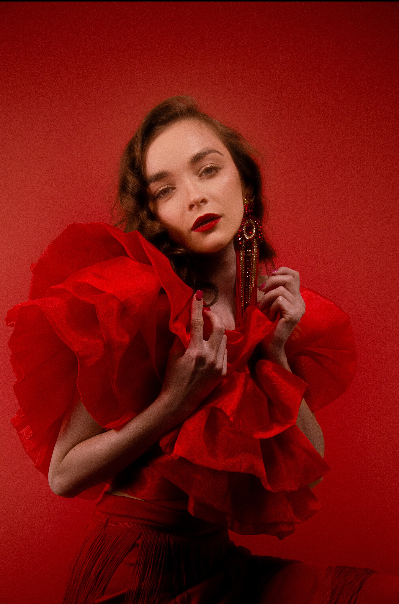 Portret kobiety w czerwonej, ekstrawaganckiej kreacji z falbanami i długimi, bogato zdobionymi kolczykami na tle jednolitego, czerwonego tła.