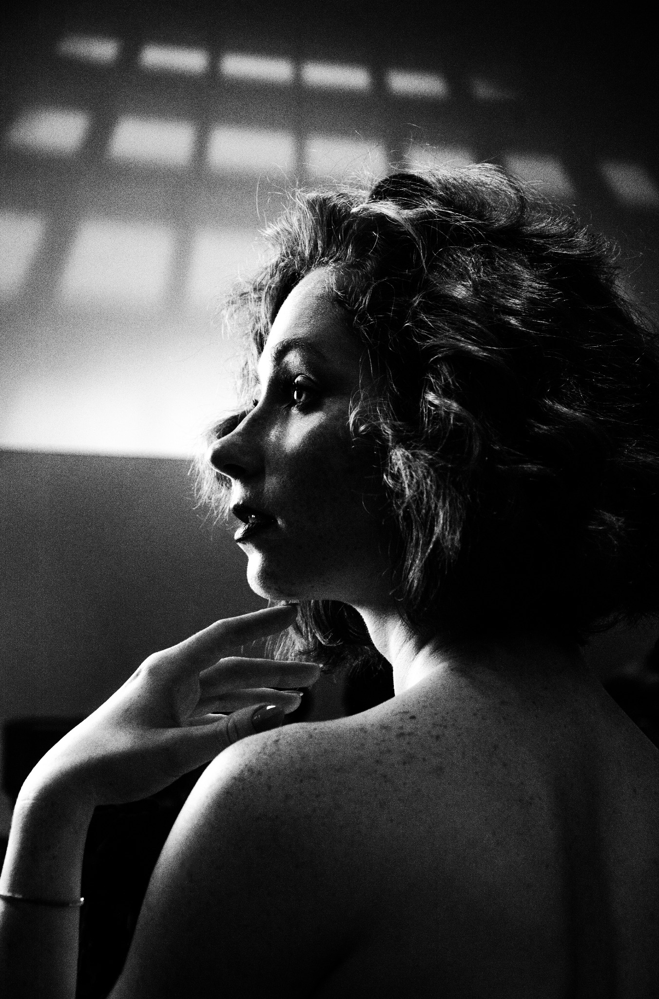 Czarno-biały portret studyjny kobiety z kręconymi włosami, profil, z ręką pod brodą, widoczne piegi na ramionach, oświetlenie padające z góry tworzące geometryczne cienie na ścianie w tle.