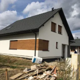 Nowoczesny dom jednorodzinny z białą elewacją i drewnianymi akcentami na oknach, widoczny stos materiałów budowlanych przed budynkiem i częściowo odsłonięta podstawa elewacji.