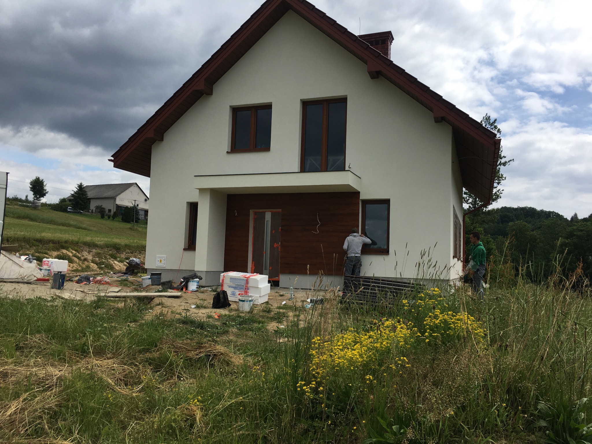 Nowo otynkowany dom jednorodzinny z brązowymi ramami okiennymi i wykończeniem elewacji drewnem, w trakcie prac wykończeniowych, z widocznymi materiałami budowlanymi i dwoma pracownikami na zewnątrz.