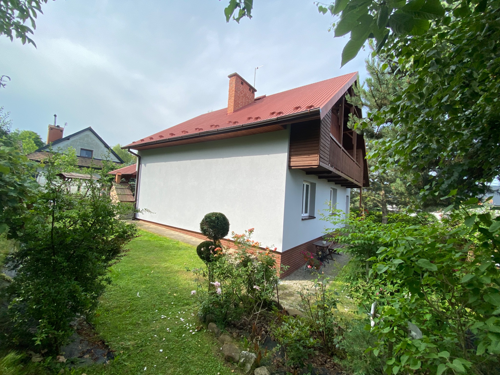 Dom jednorodzinny z jasną elewacją, czerwoną blachodachówką i drewnianą werandą, otoczony zielenią ogrodu z ozdobnymi krzewami.