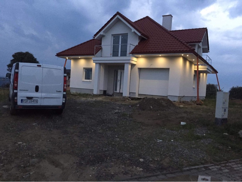 Nowoczesny dom jednorodzinny z białymi ścianami i czerwoną dachówką, oświetlony punktowymi lampami elewacyjnymi, z zaparkowanym białym vanem Renault z tyłu, na tle pochmurnego nieba.