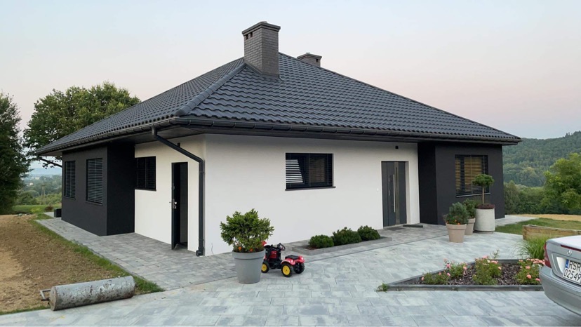 Nowoczesny dom jednorodzinny z białą elewacją i czarnymi akcentami, ciemna dachówka, szara kostka brukowa na podjeździe, w tle zalesione wzgórza.