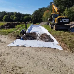 Przygotowanie podłoża pod drogę: rozłożona geowłóknina, na niej pryzmy żwiru, w tle koparka JCB i pracownicy.