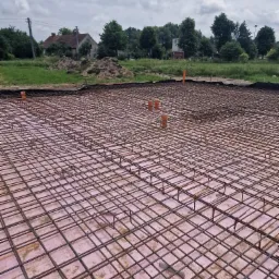 Siatka zbrojeniowa ułożona na różowym styropianie, przygotowana do wylewki fundamentów, widoczne pionowe rury instalacyjne