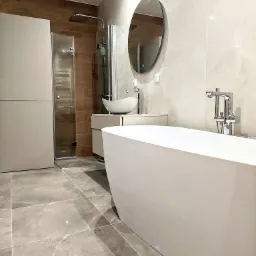 Nowoczesna łazienka z wolnostojącą białą wanną, owalnym lustrem z podświetleniem LED, kabiną prysznicową typu walk-in i szarymi płytkami podłogowymi imitującymi marmur.