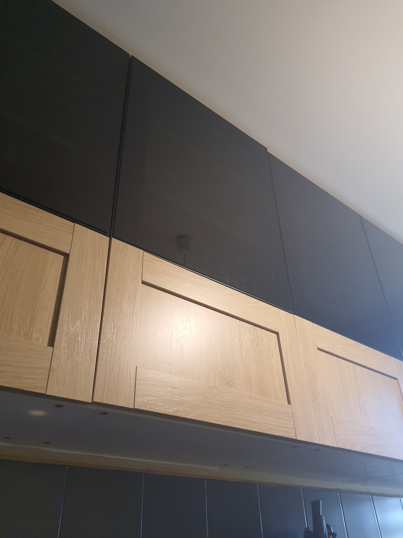 Nowoczesne szafki kuchenne: dolne z jasnego drewna z frezowanymi frontami, górne z czarnego szkła, widok od dołu.