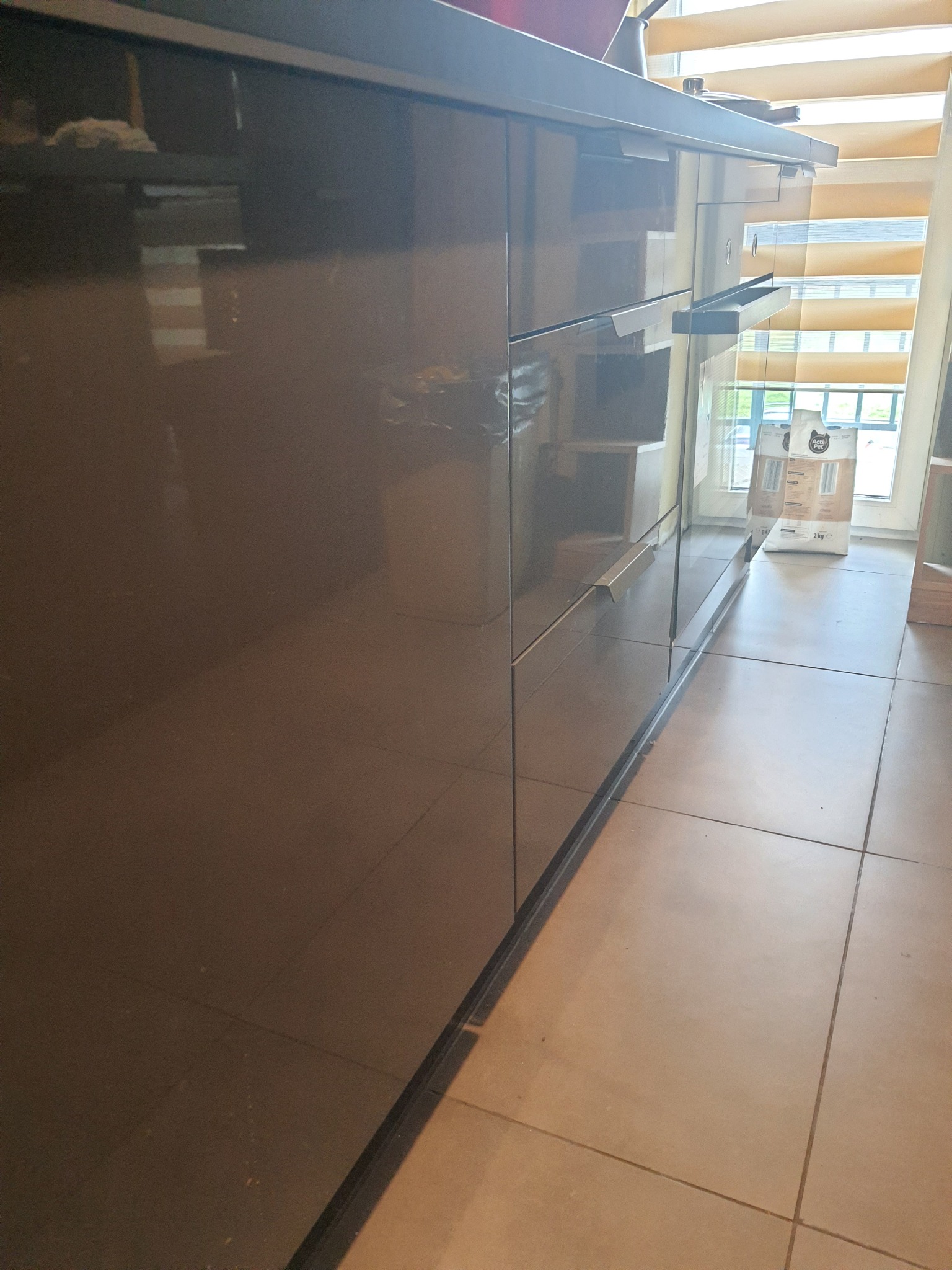 Błyszcząca, ciemnobrązowa zabudowa kuchenna z metalowymi uchwytami, widok z dołu, na podłodze płytki, w tle okno z żaluzjami.