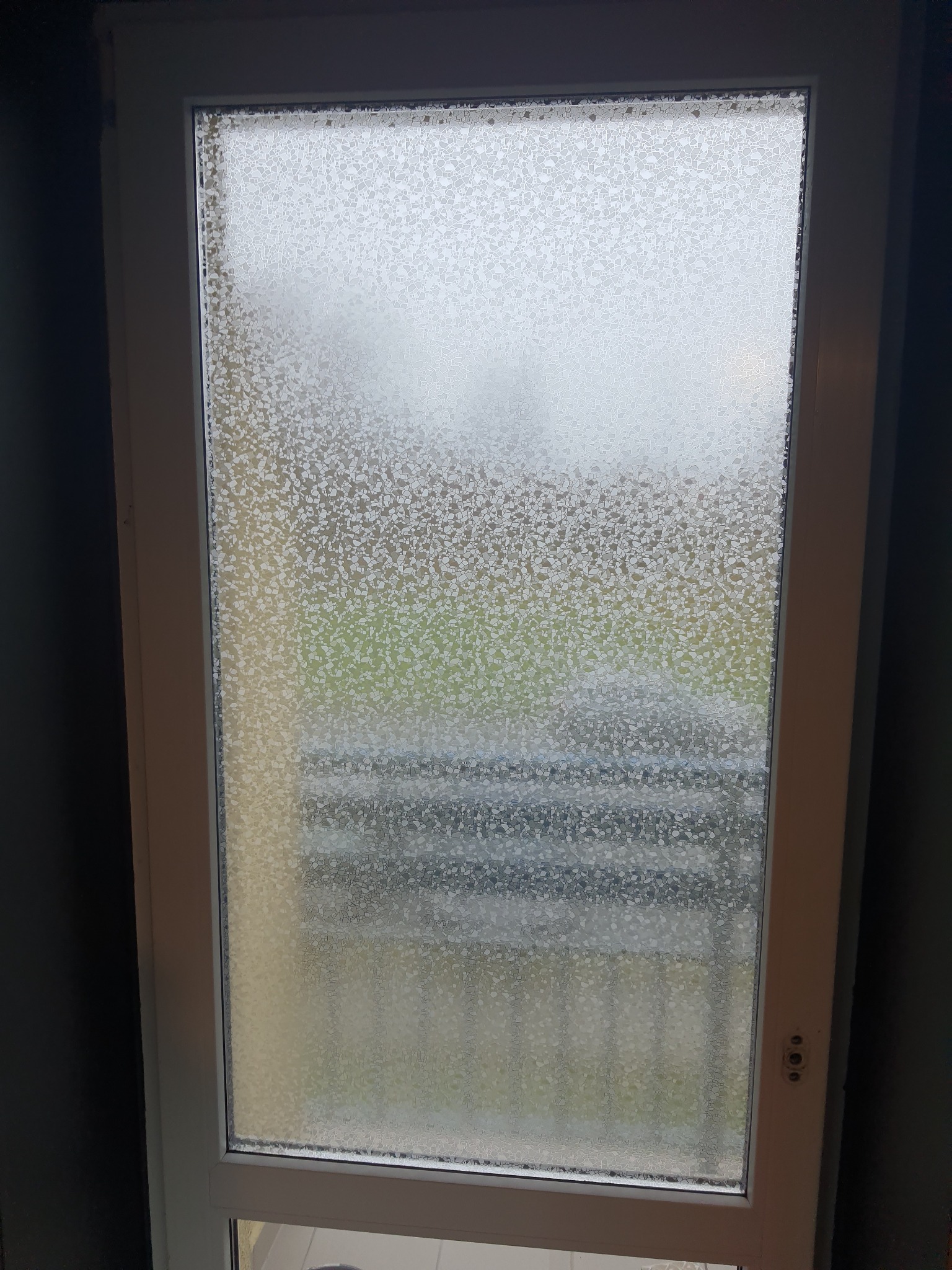 Biała rama okienna z szybą o fakturze imitującej oszronienie, przez którą widać zamazany widok zieleni i szarości na zewnątrz.