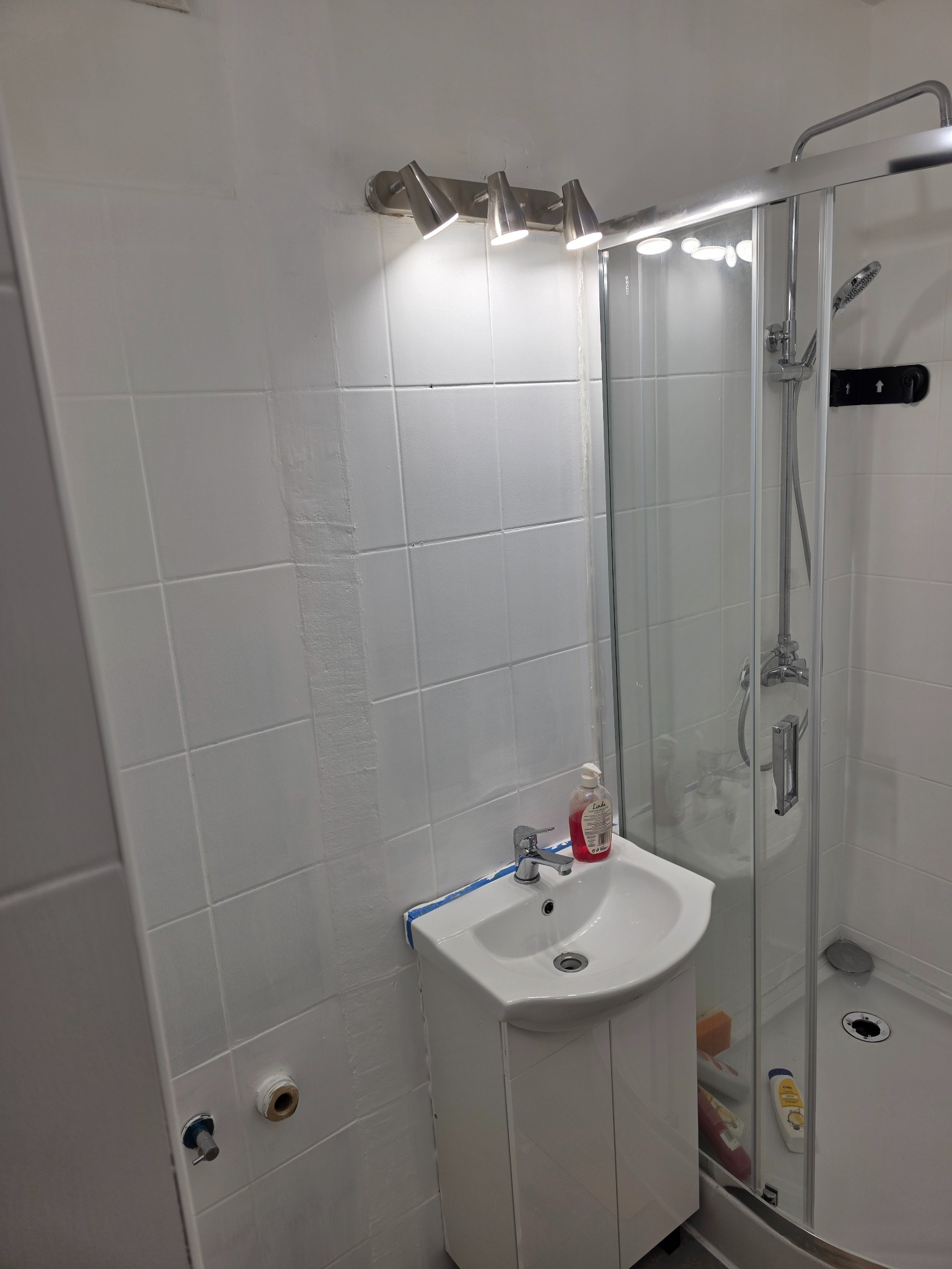 Biała łazienka po malowaniu, z kabiną prysznicową i umywalką. Widoczne świeże białe ściany i drobne niedociągnięcia w wykończeniu.