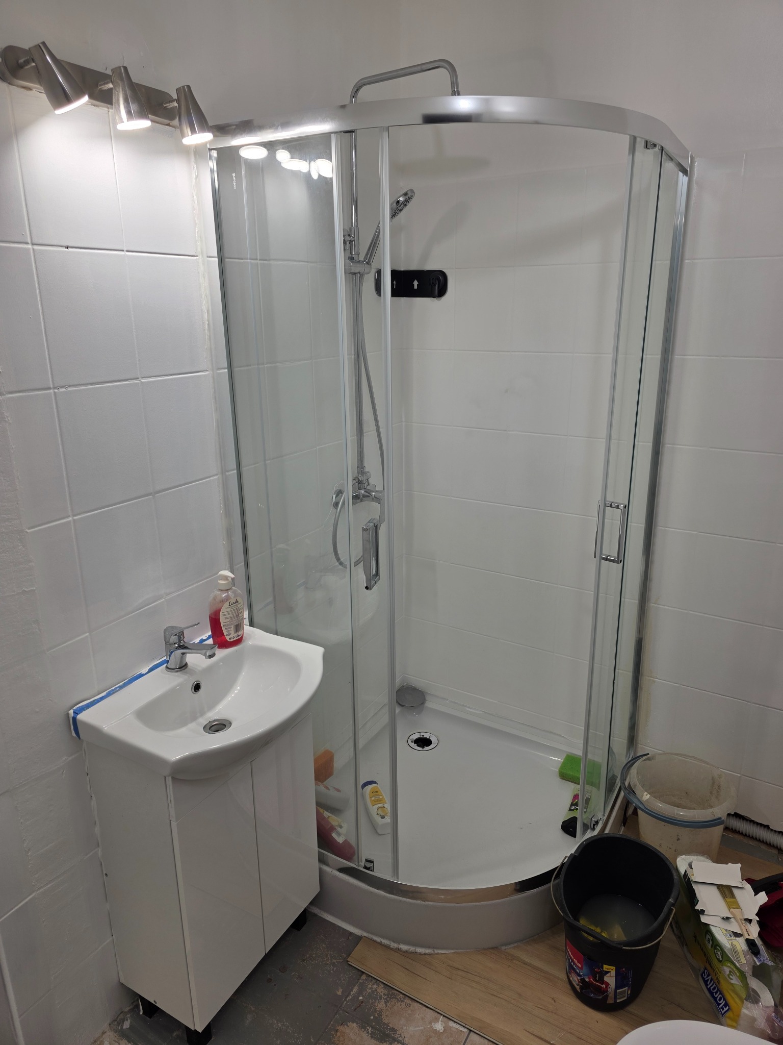 Biała kabina prysznicowa z umywalką w narożnej, wykafelkowanej łazience. Widoczne narzędzia i akcesoria remontowe na podłodze.