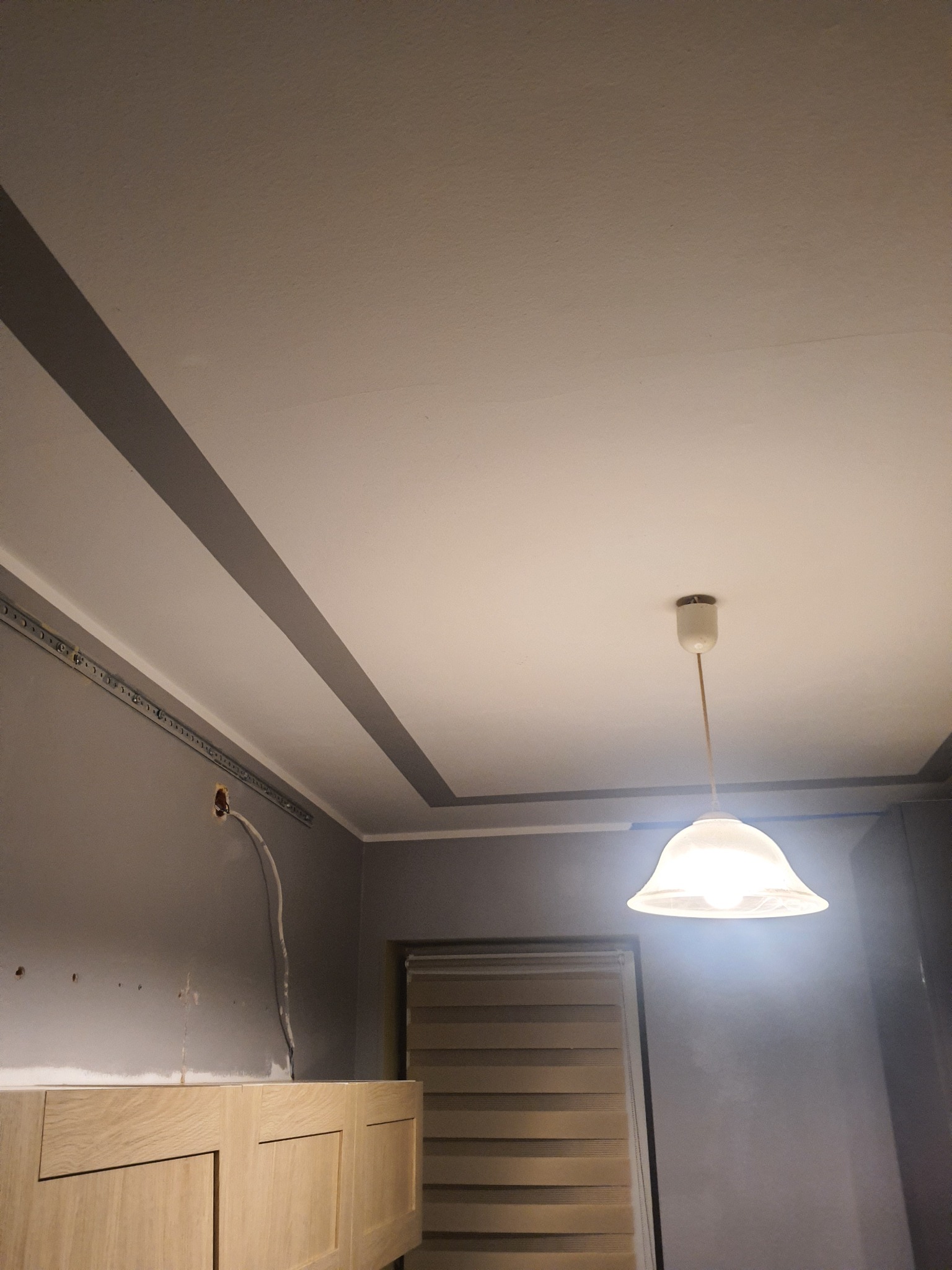 Szare ściany po malowaniu z widocznymi przewodami elektrycznymi, biały sufit z dekoracyjnymi pasami i wiszącą lampą, fragment zabudowy meblowej z jasnego drewna.