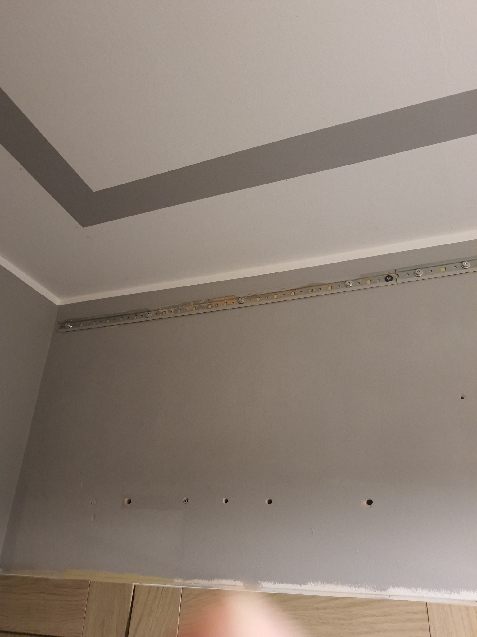 Szara ściana z zamontowaną metalową szyną montażową pod sufitem, widoczne otwory w ścianie i ślady po szpachlowaniu.