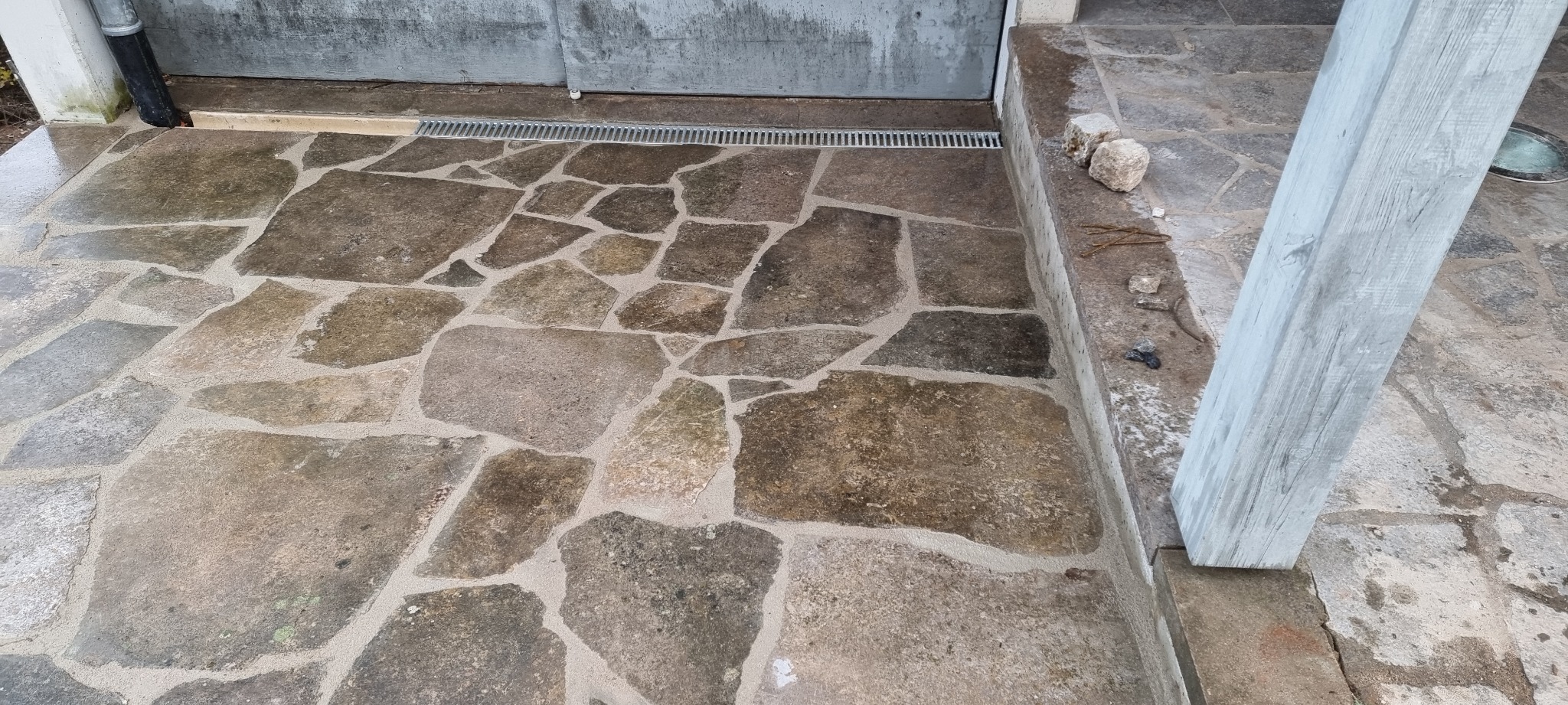 Ułożona z nieregularnych kamieni posadzka na zewnątrz budynku, z widoczną kratką odpływową i fragmentem drewnianej kolumny pomalowanej na szaro.