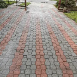 Mokra kostka brukowa ułożona w pasy z szarych i czerwonych elementów, prowadząca w stronę żelaznej bramy i ulicy.