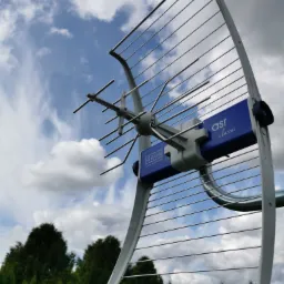 Antena telewizyjna typu Yagi-Uda z niebieskim elementem centralnym marki ASR Classic, skierowana ku niebu z chmurami i zielonymi drzewami w tle.