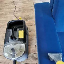 Czyszczenie niebieskiego narożnika za pomocą odkurzacza piorącego Karcher, widoczne ślady wilgoci na tapicerce, parkiet w tle.