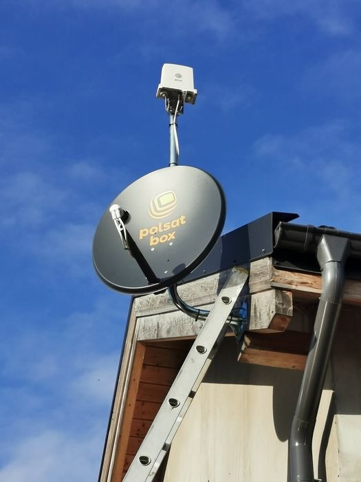 Antena satelitarna Polsat Box zamontowana na dachu budynku, widoczna drabina oparta o ścianę, błękitne niebo w tle. Instalacja antenowa.