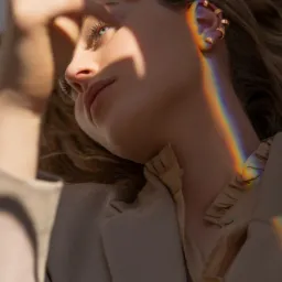 Portret kobiety w beżowym żakiecie, z tęczowym refleksem na twarzy i złotymi kolczykami na uchu, dłoń z pierścionkiem w górnym lewym rogu kadru.