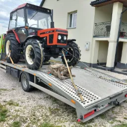Pomarańczowy traktor Zetor 7245 zabezpieczony pasami na platformie lawety transportowej, na tle jasnego domu z balkonem i zielonego pola.