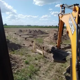 Żółta koparka JCB przy wykopie pod fundamenty z drewnianym szalunkiem, widok z kabiny, w tle pole i osoba nadzorująca.
