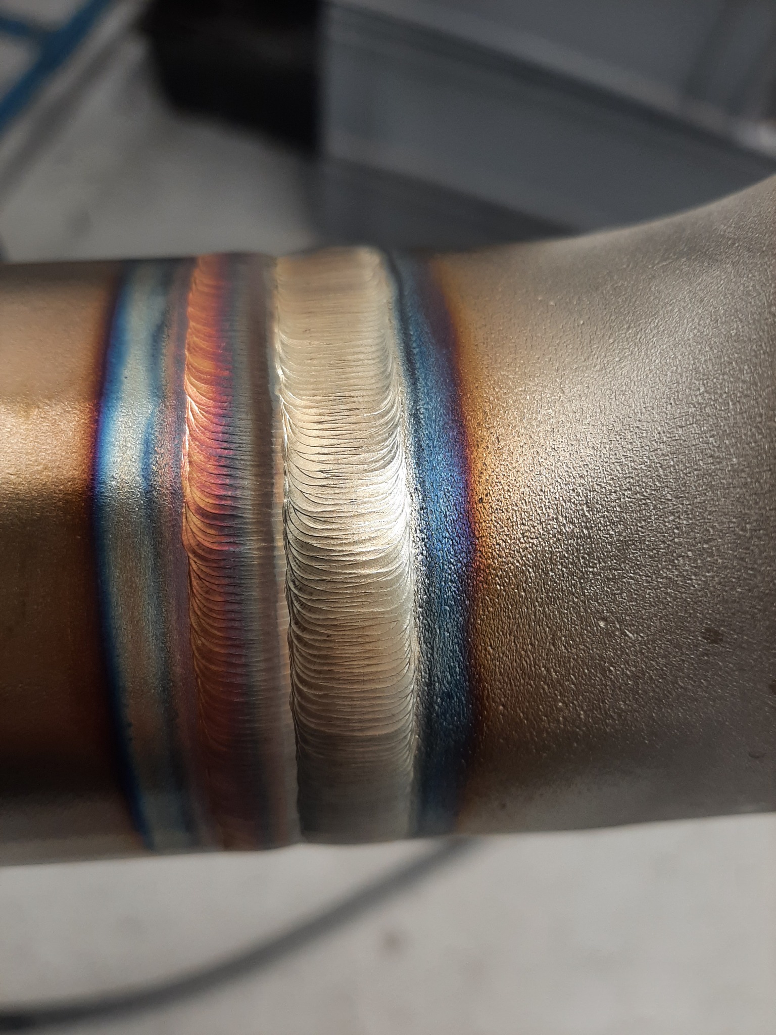 Szczegółowe zbliżenie spoiny na rurze ze stali nierdzewnej, widoczne charakterystyczne przebarwienia termiczne w odcieniach niebieskiego, fioletowego i złotego, świadczące o precyzyjnym procesie...