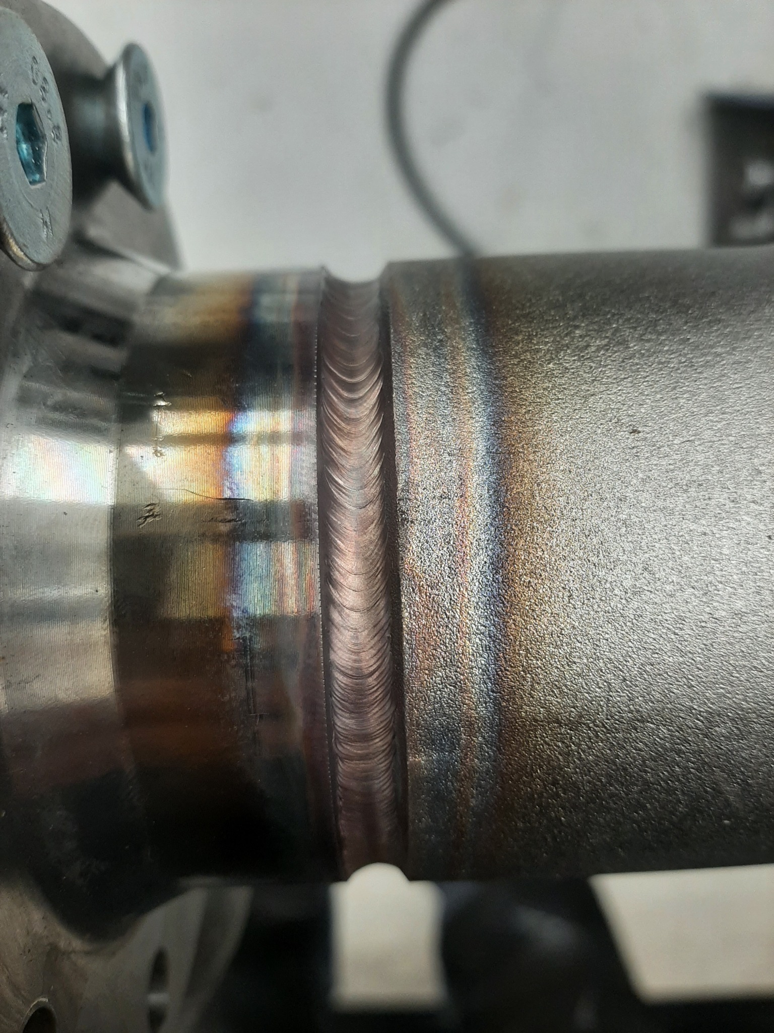 Szczegółowe zbliżenie na okrągły, metalowy element z widocznym, precyzyjnym spawem, prezentującym charakterystyczne tęczowe zabarwienia termiczne na powierzchni.