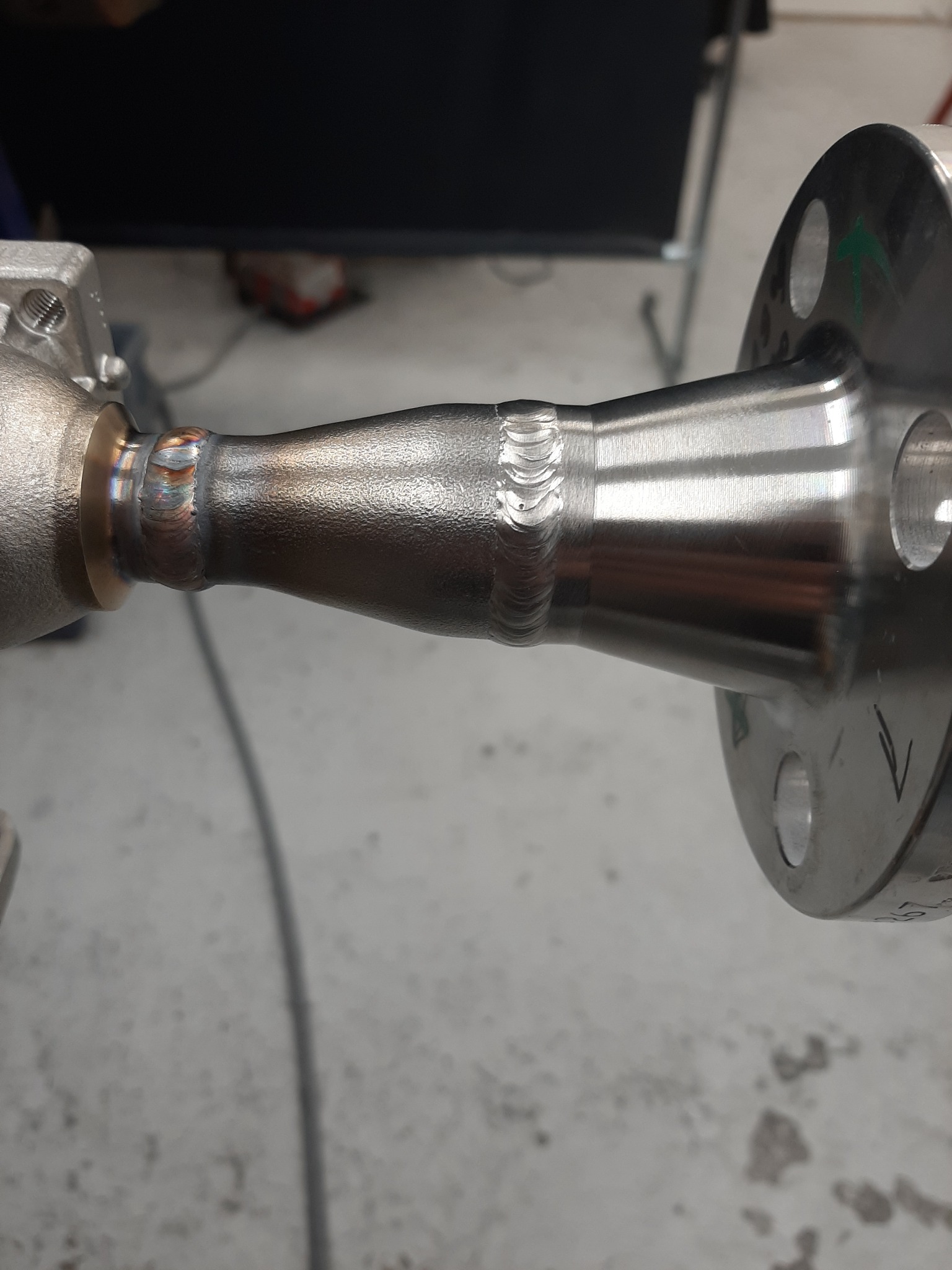 Precyzyjnie wykonane spawy łączące metalowe elementy, widoczne ślady po procesie spawania na powierzchni metalu, strzałka zaznaczona zielonym markerem na kołnierzu.