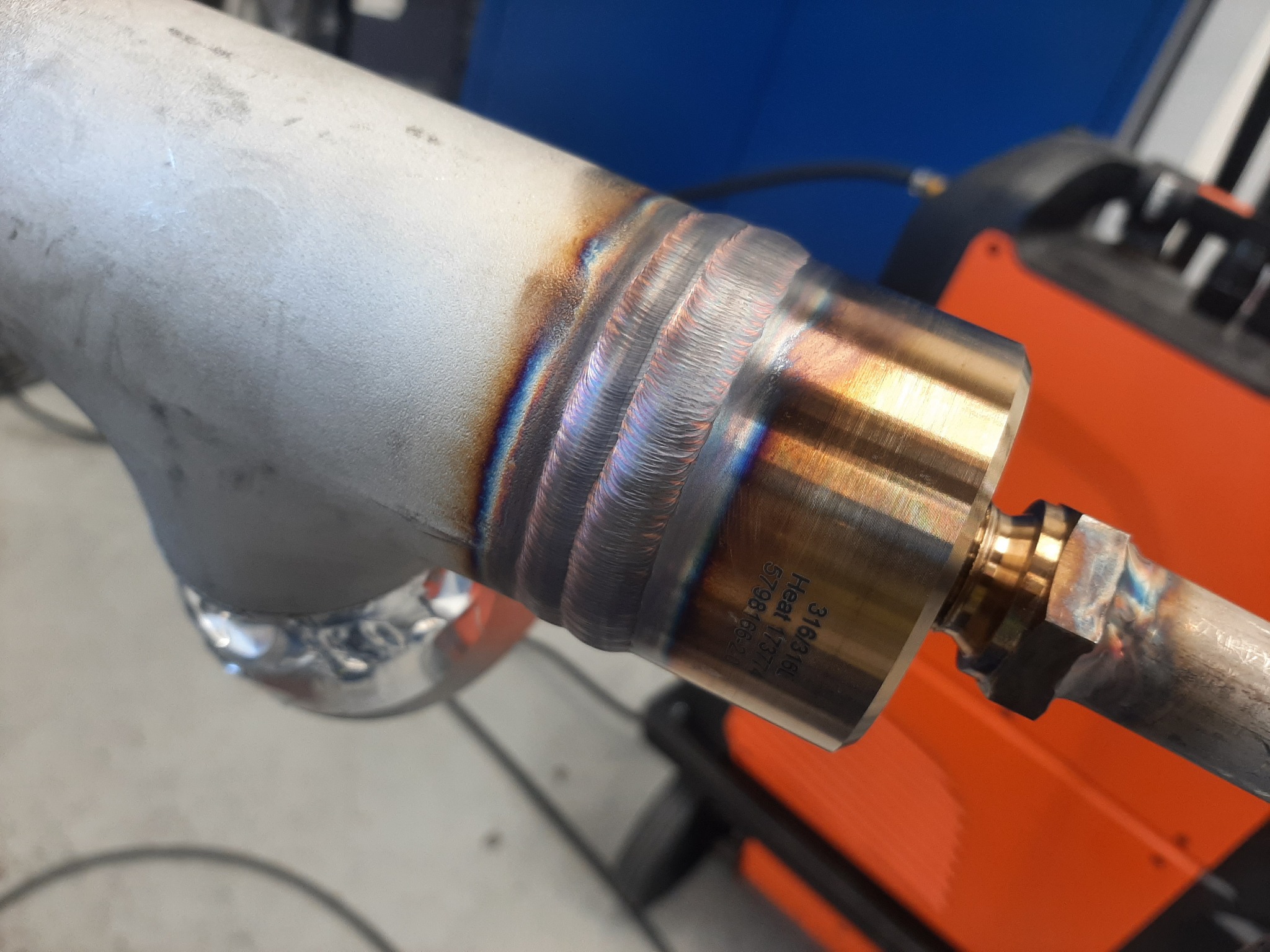 Spoina TIG łącząca rurę ze złączką, widoczne ślady ciepła i precyzyjne spawy, w tle pomarańczowa spawarka.