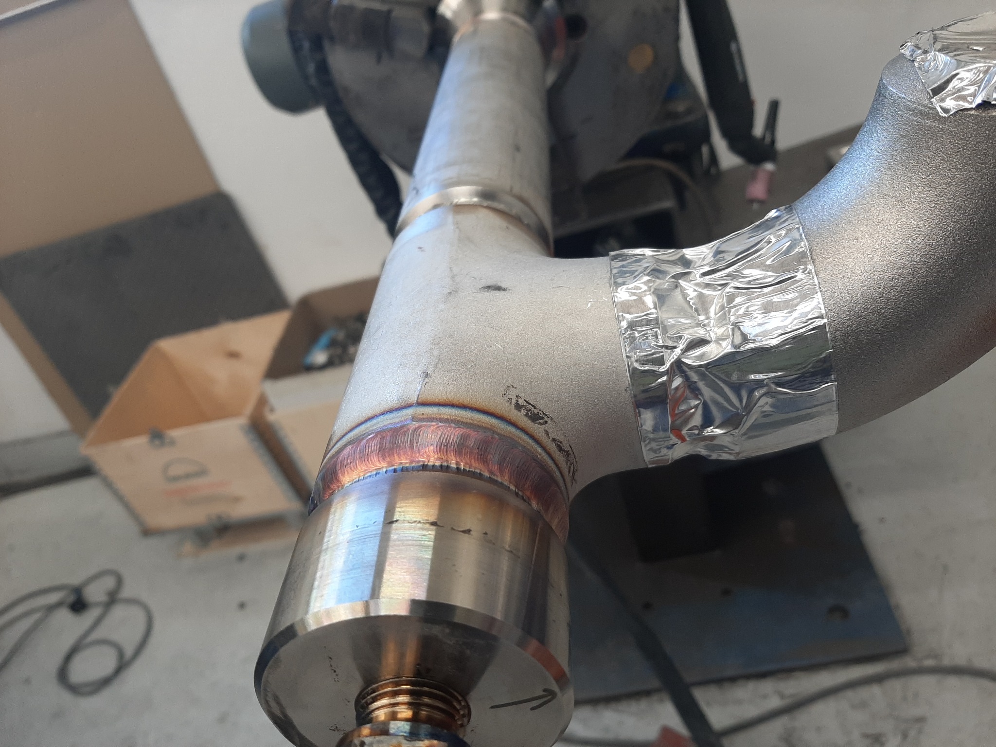 Połączenie spawane rur ze stali nierdzewnej, owinięte folią aluminiową, widoczne ślady spawania TIG, w tle skrzynka narzędziowa.