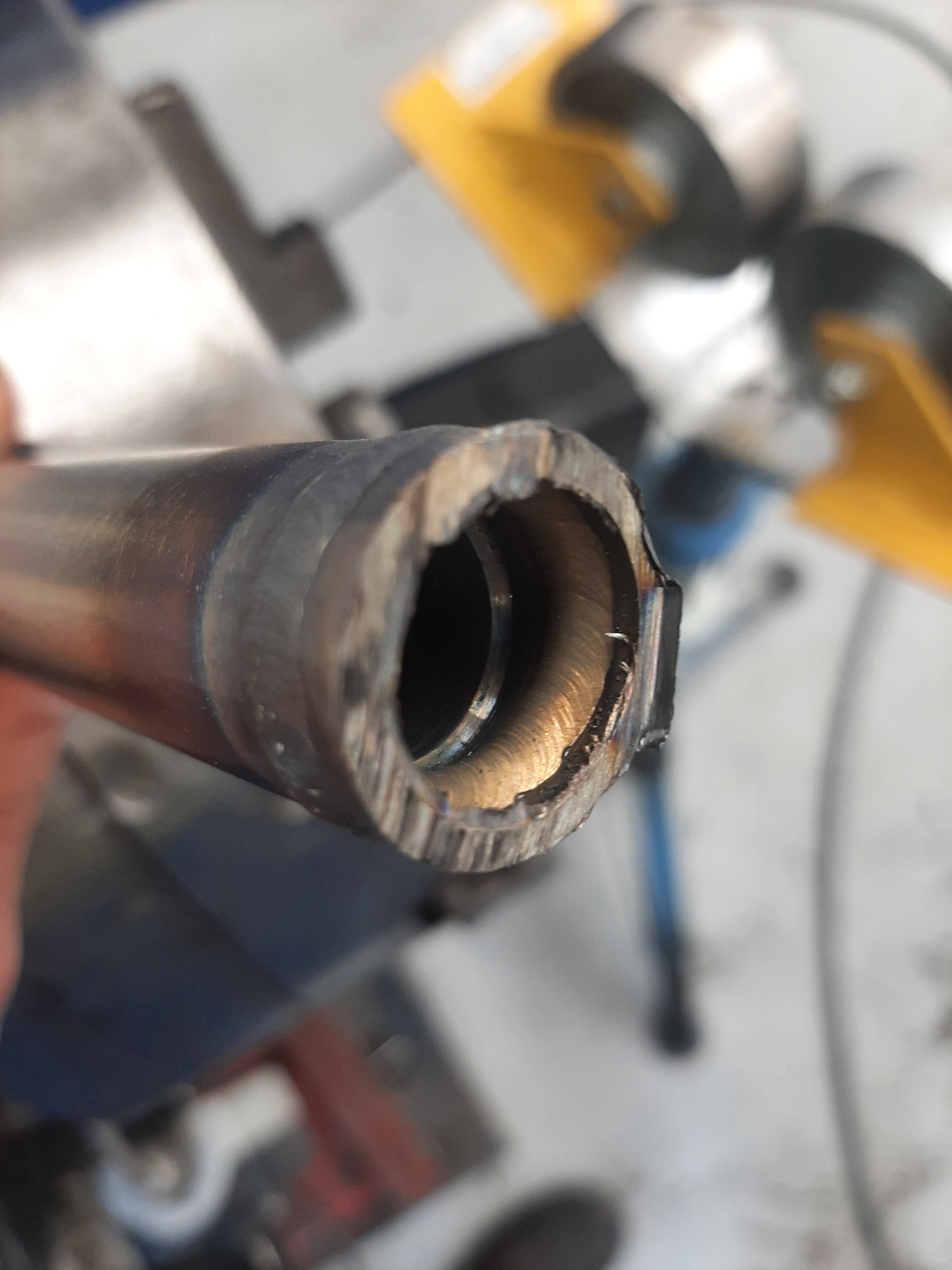 Metalowa rura z widocznymi śladami obróbki, nierównymi krawędziami i wewnętrznymi rowkami, uchwycona z bliska pod kątem, z elementami warsztatu w tle.