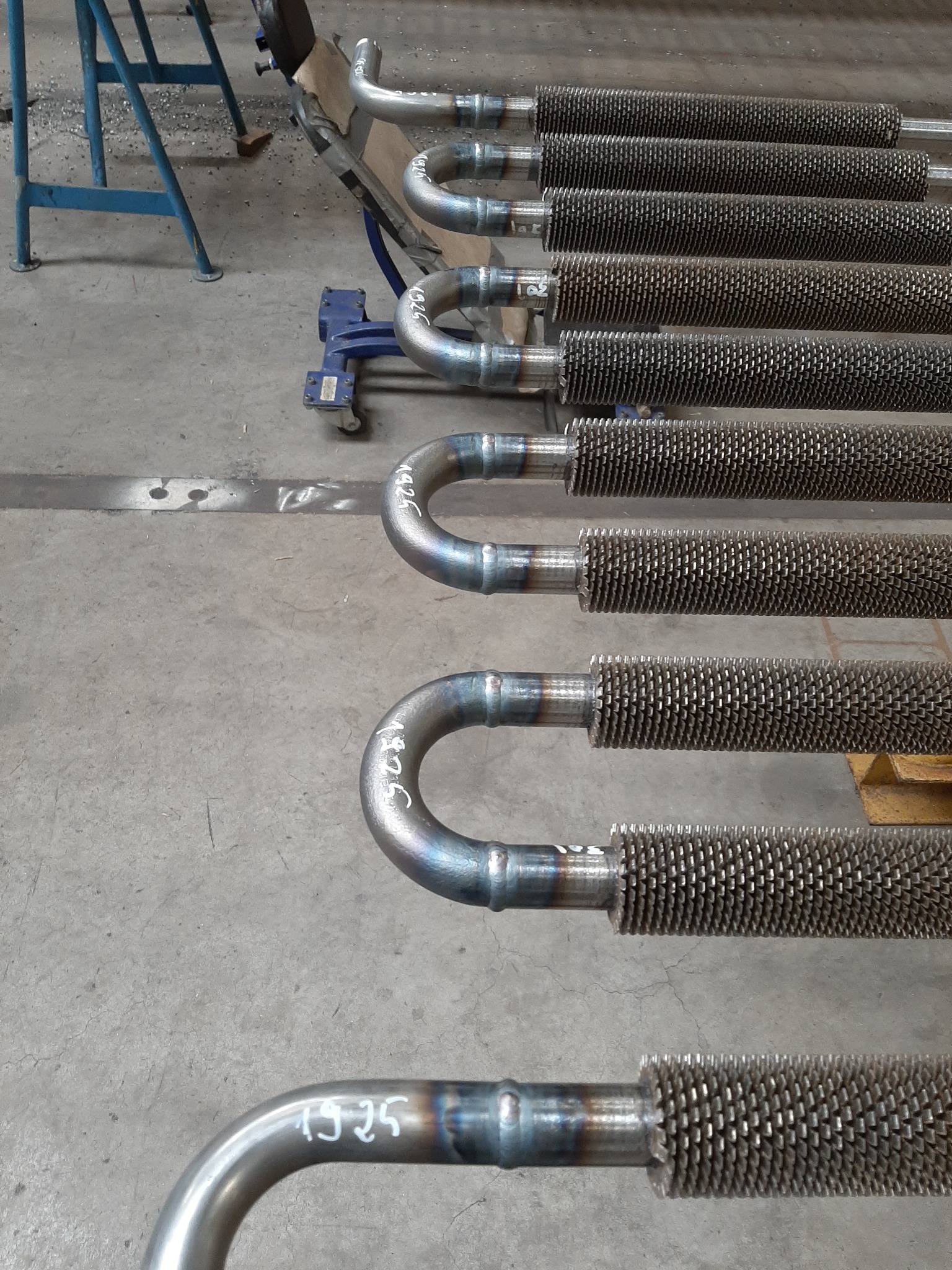 Rzędy rur z żebrowaniem, połączone spawami, leżące na betonowej posadzce; na jednej z rur widoczny napis '1925'.