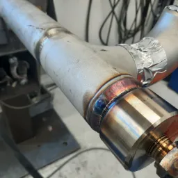 Połączenie spawane rury ze stali nierdzewnej, z widocznymi śladami spawania TIG i zabezpieczeniem termicznym w postaci folii aluminiowej na zagięciu.