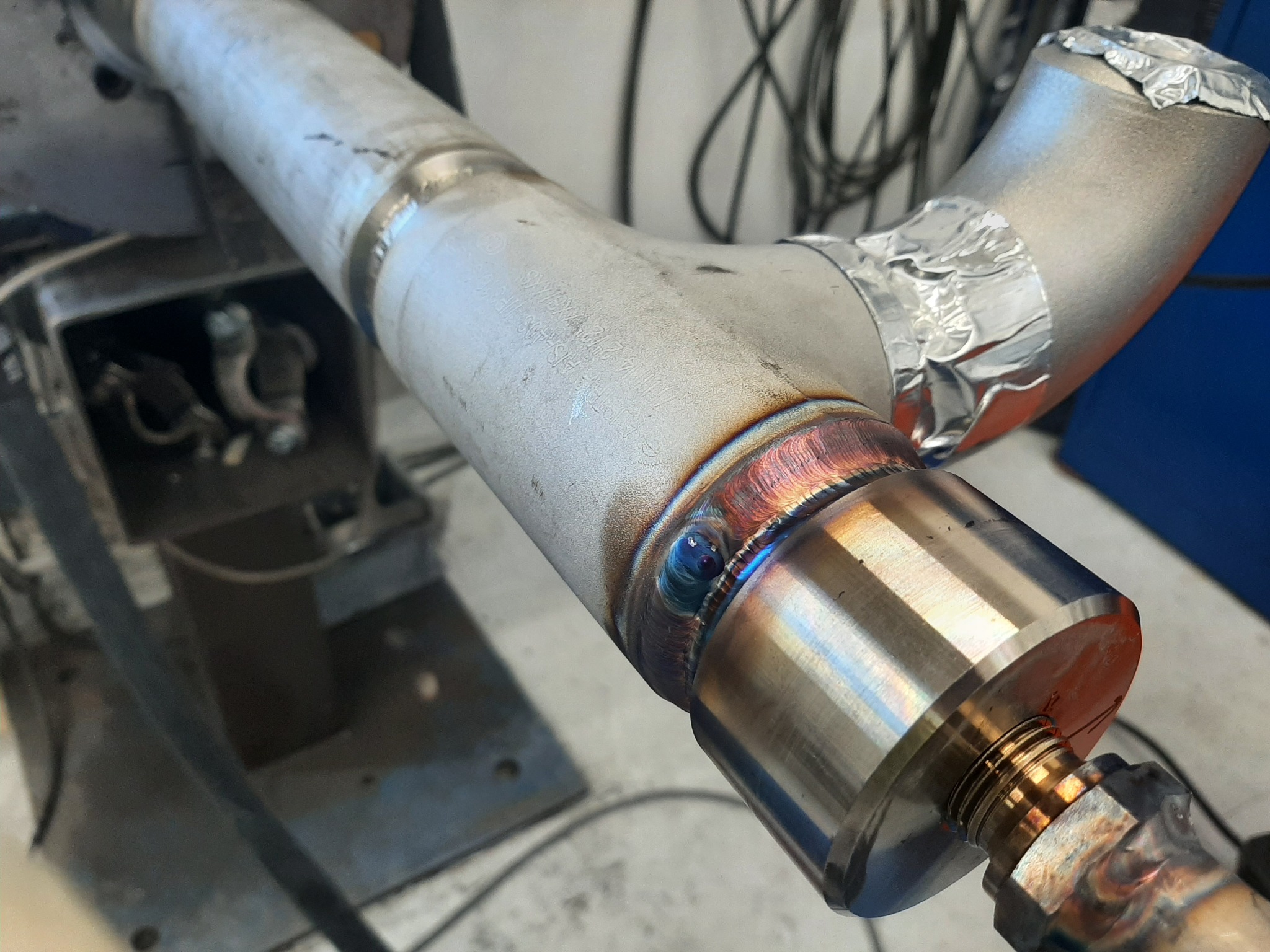 Połączenie spawane rury ze stali nierdzewnej, z widocznymi śladami spawania TIG i zabezpieczeniem termicznym w postaci folii aluminiowej na zagięciu.